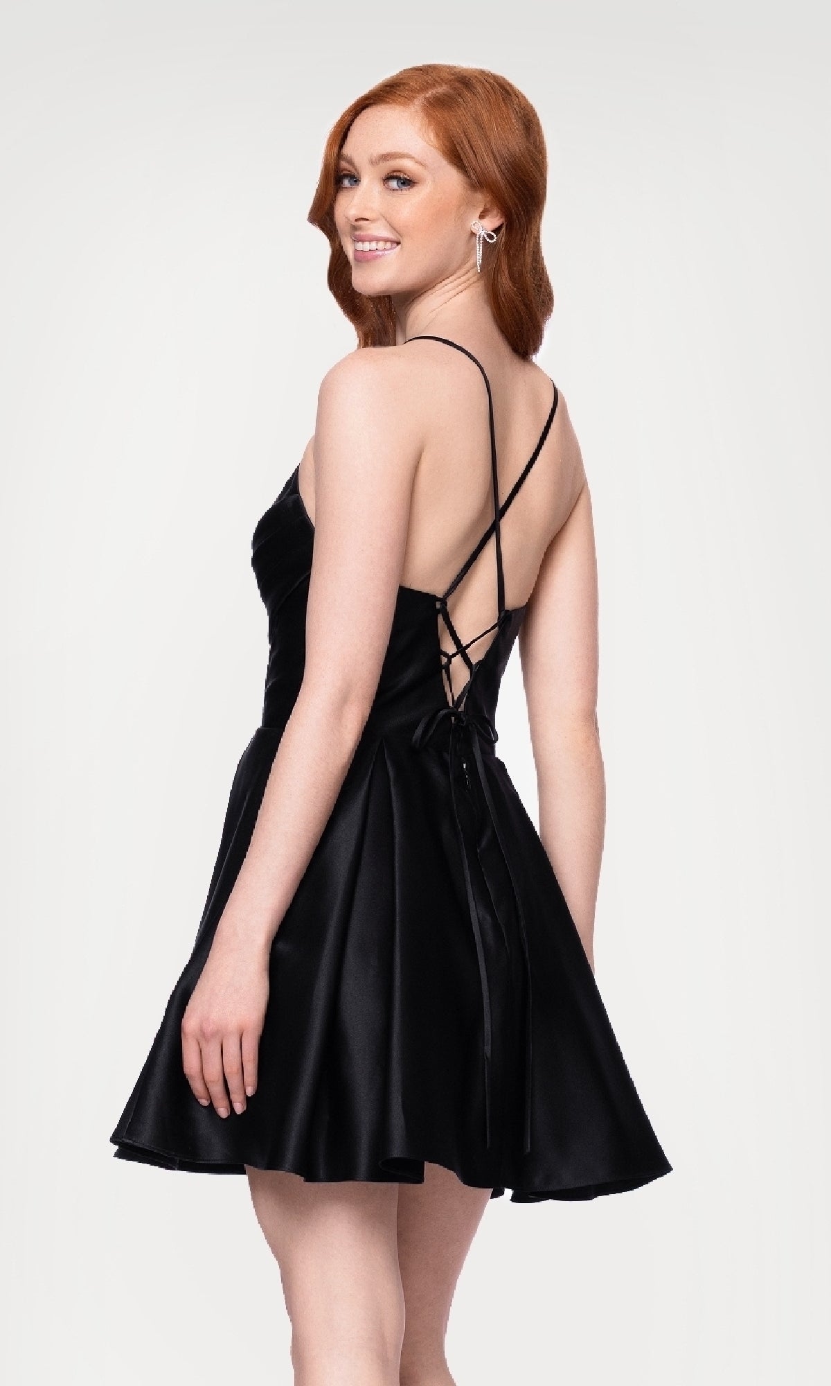Woman models the Lace-Up Short Hoco Dress: Blondie Nites 4578BN—a sleeveless black junior cocktail dress with a flared skirt and crisscross back straps—smiling over her shoulder against a plain light background.